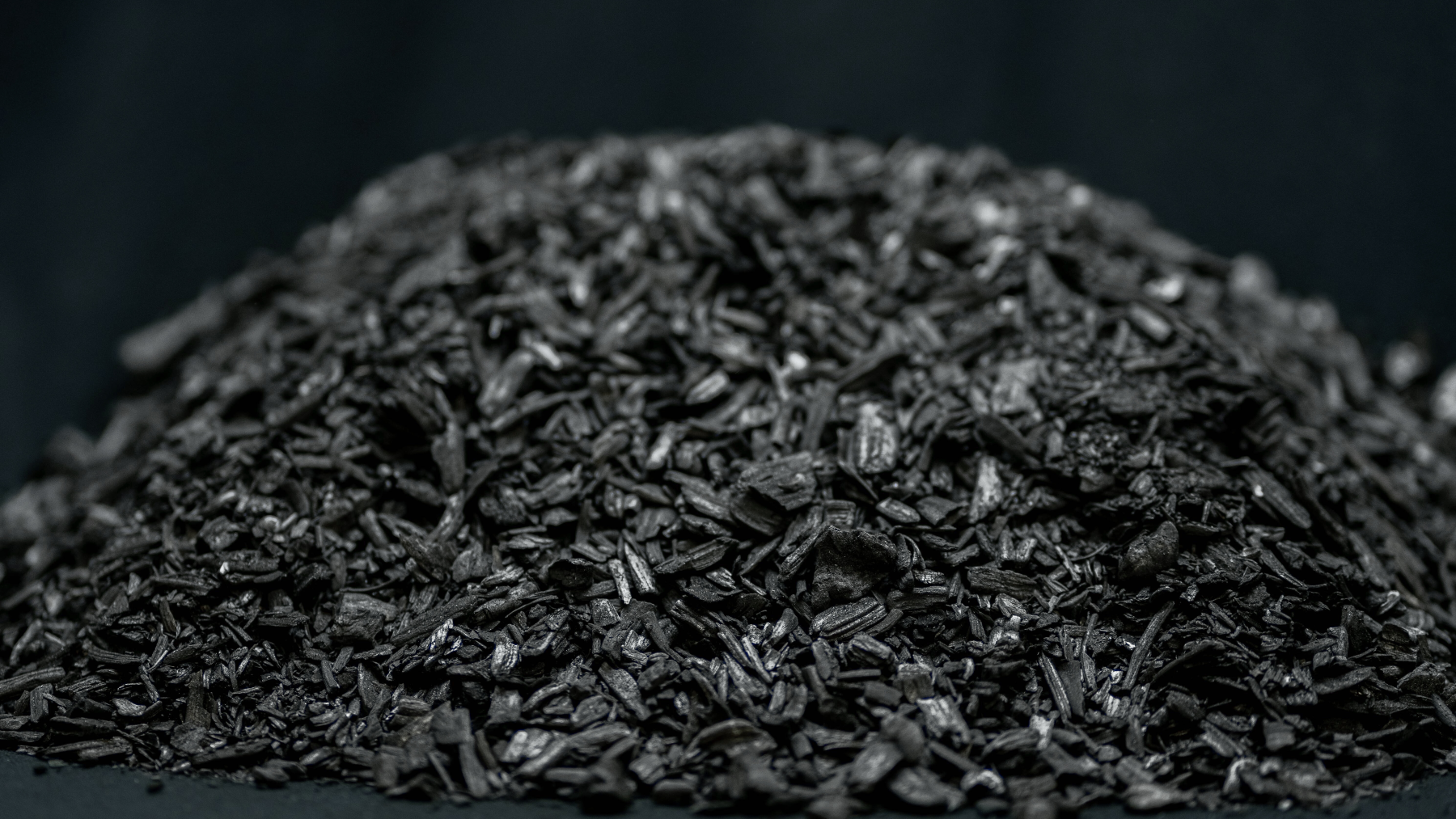 Die Nahaufnahme zeigt einen dunklen Haufen grobkörniger Pflanzenkohle auf schwarzem Hintergrund. Die einzelnen Stücke der Kohle sind unregelmäßig geformt, kantig und haben eine matte bis leicht glänzende Oberfläche. Die Textur wirkt faserig und erinnert an verkohltes Holz oder Rindenreste. Dieses Material stellt TerraCoal da – eine spezielle Form der Pflanzenkohle, die zur Bodenverbesserung und CO₂-Bindung eingesetzt wird.